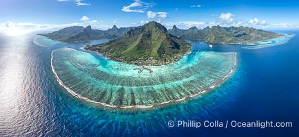 Barrier Reef around Moorea Island, French Polynesia. Cook's Bay to the left, Opunohu Bay to the right. The outer reef slope is seen adjacent to deep blue oceanic water with white waves breaking against the reef edge. Next, a wide shallow reef flat occurs dotted with coral bommies. Inside of that, a shallow protected lagoon is formed against the island.  Tall, rugged, eroded mountains are seen hinting at the age of the ancient volcano that originally formed the island and that is now sinking back down, leaving the encircling reef behind