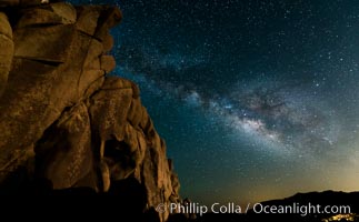The Milky Way rises above a huge wall of stone, stars fill the night sky and soar over the distant lights of campers, Joshua Tree National Park, California