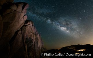 The Milky Way rises above a huge wall of stone, stars fill the night sky and soar over the distant lights of campers, Joshua Tree National Park, California