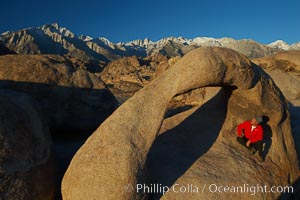 A hiker admires Mobius Arch in early morning golden sunlight, with the snow-covered Sierra Nevada Range and the Alabama Hills seen in the background, Alabama Hills Recreational Area