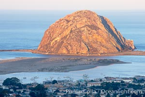 Morro Rock lit at sunrise, rises above Morro Bay which is still in early morning shadow